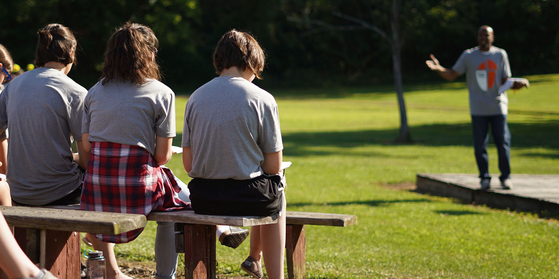 Three campers sit on a bench outside listening to man preach.